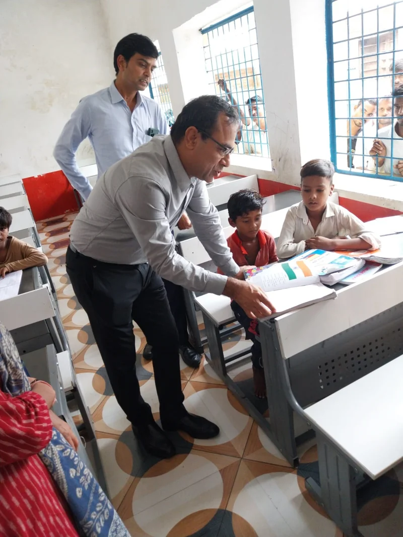 सरकारी स्कूल की कक्षा में मेज पर खुली किताब देखते अधिकारी और विद्यार्थी